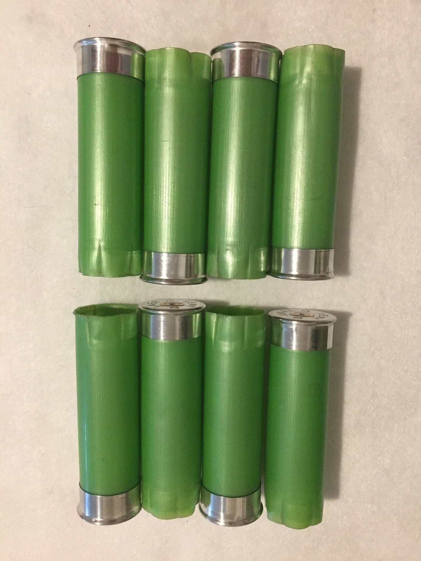 8 empty lime green 12 gauge shotgun shells with silver bases arranged in 2 rows on a white surface