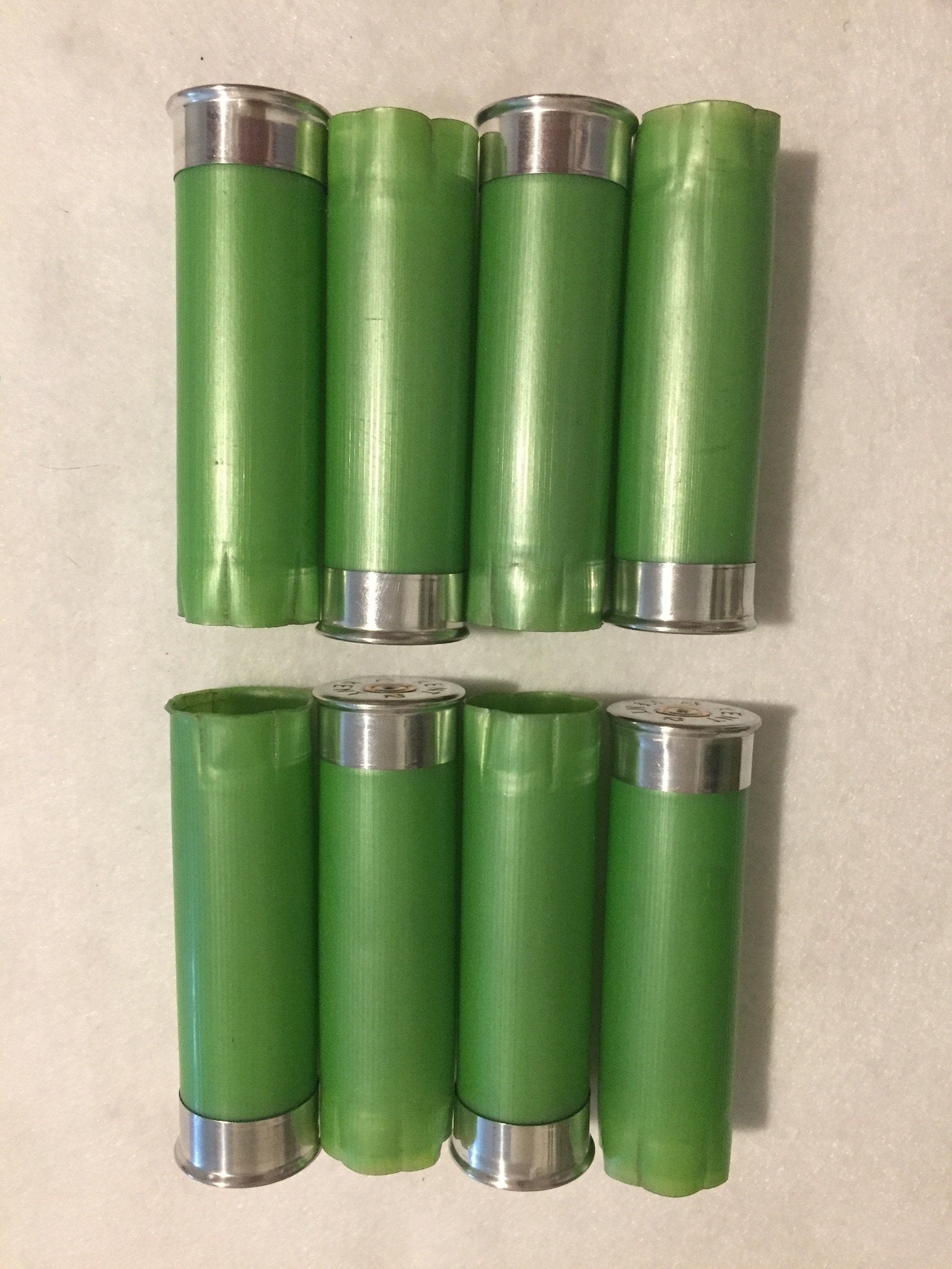 8 empty lime green 12 gauge shotgun shells with silver bases arranged in 2 rows on a white surface