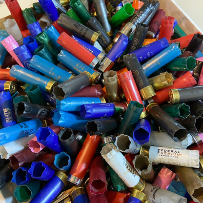 Top down view looking into a bulk pile of spent 12 gauge shotgun hulls showing open crimped plastic ends in mixed colors.