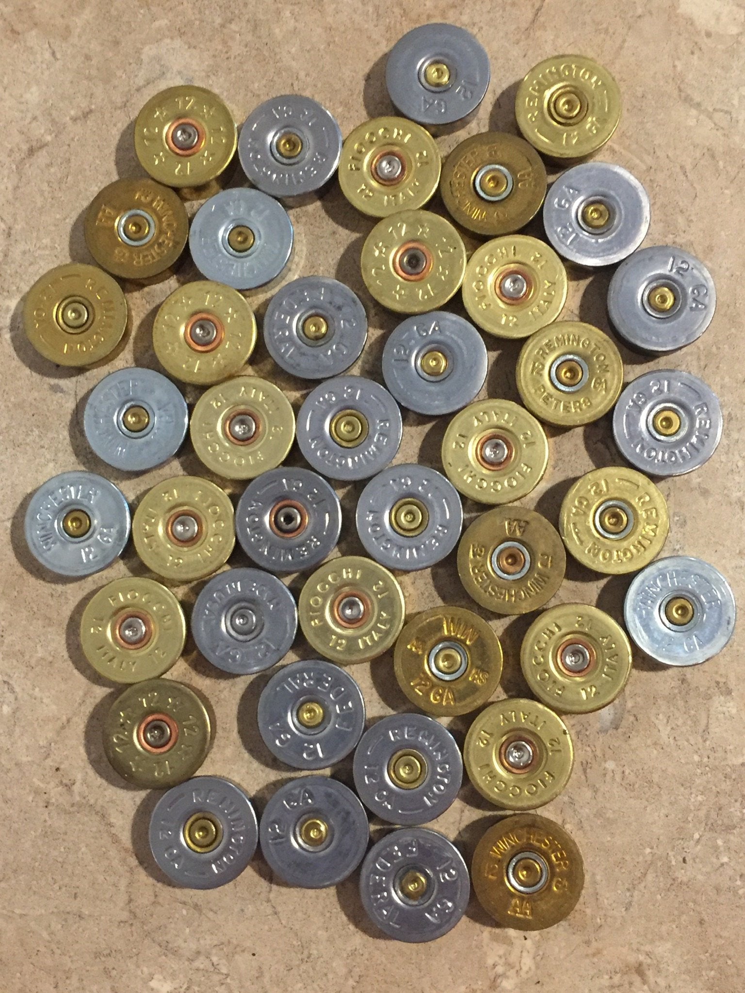 A collection of mixed gold and silver-toned shotgun shell bottoms from various brands on a textured surface.
