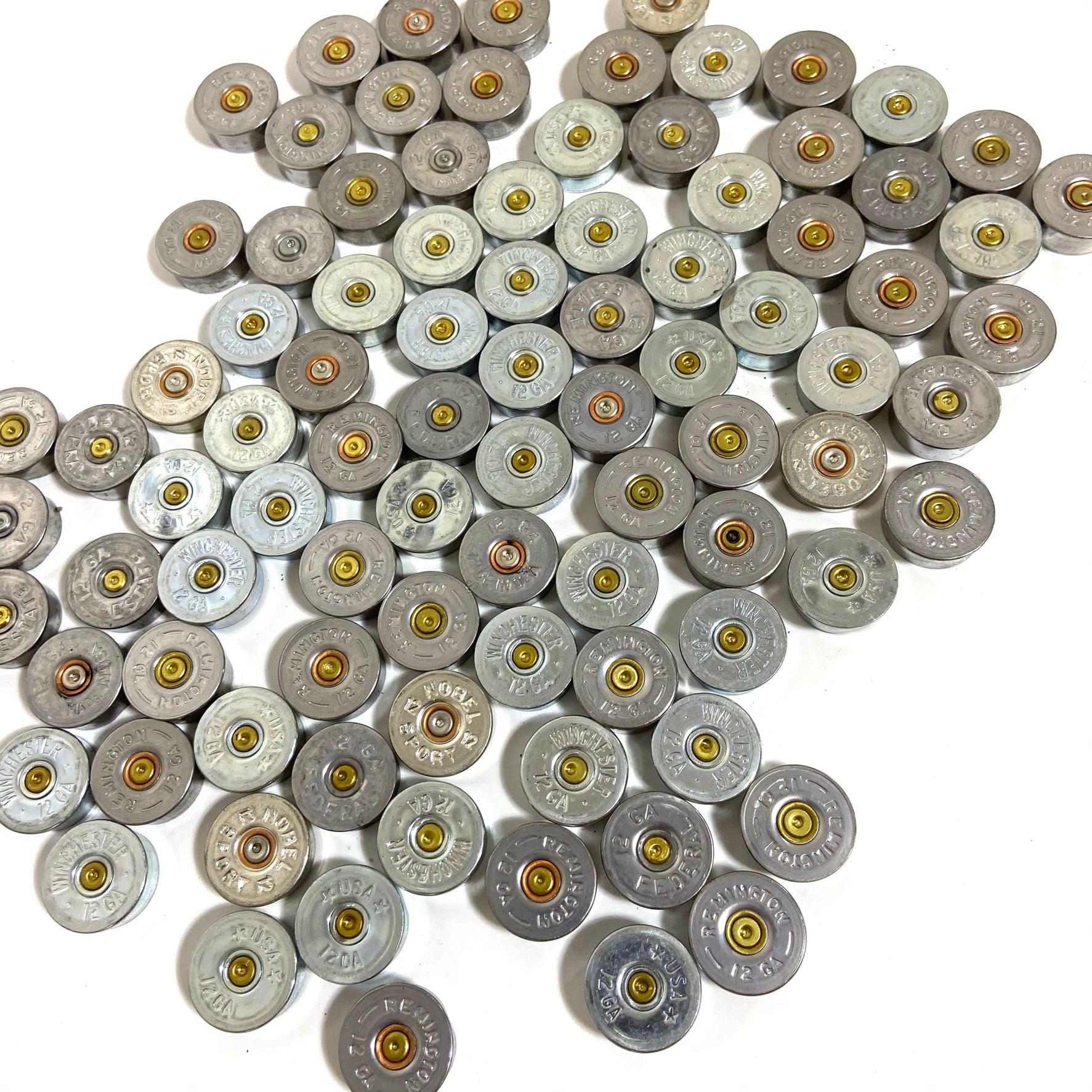 Silver 12 gauge shotgun shell bottoms scattered together on a white background, mixed brands.