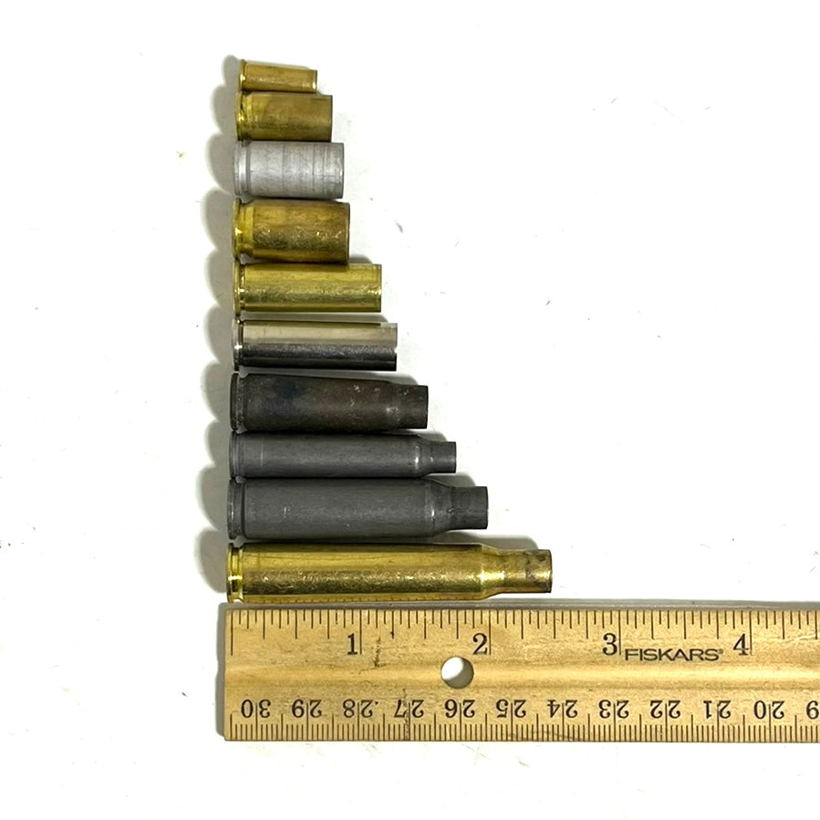 Eleven mixed spent casings in brass, nickel, and steel lined up by size next to a wooden ruler for scale.