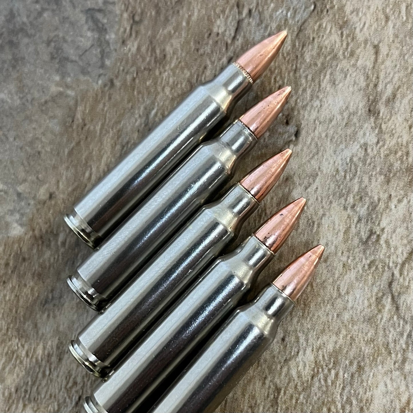 Five nickel-plated .223 dummy rifle cartridges with copper bullet tips lined up diagonally on a slate surface.