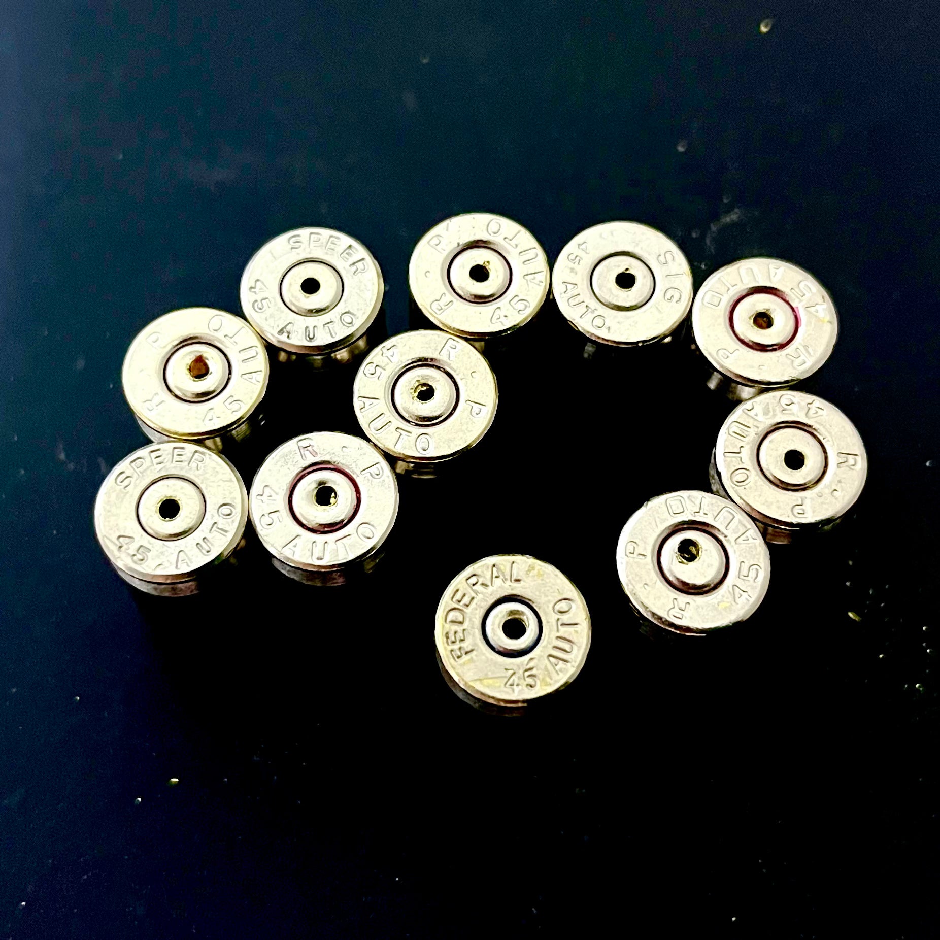 Ten nickel 45 ACP casings with drilled centers arranged in an oval showing mixed headstamps