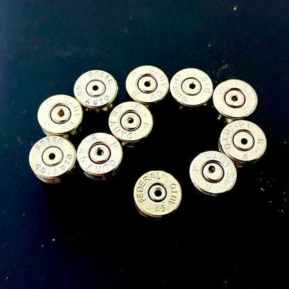 Ten nickel 45 ACP casings with drilled centers arranged in an oval showing mixed headstamps