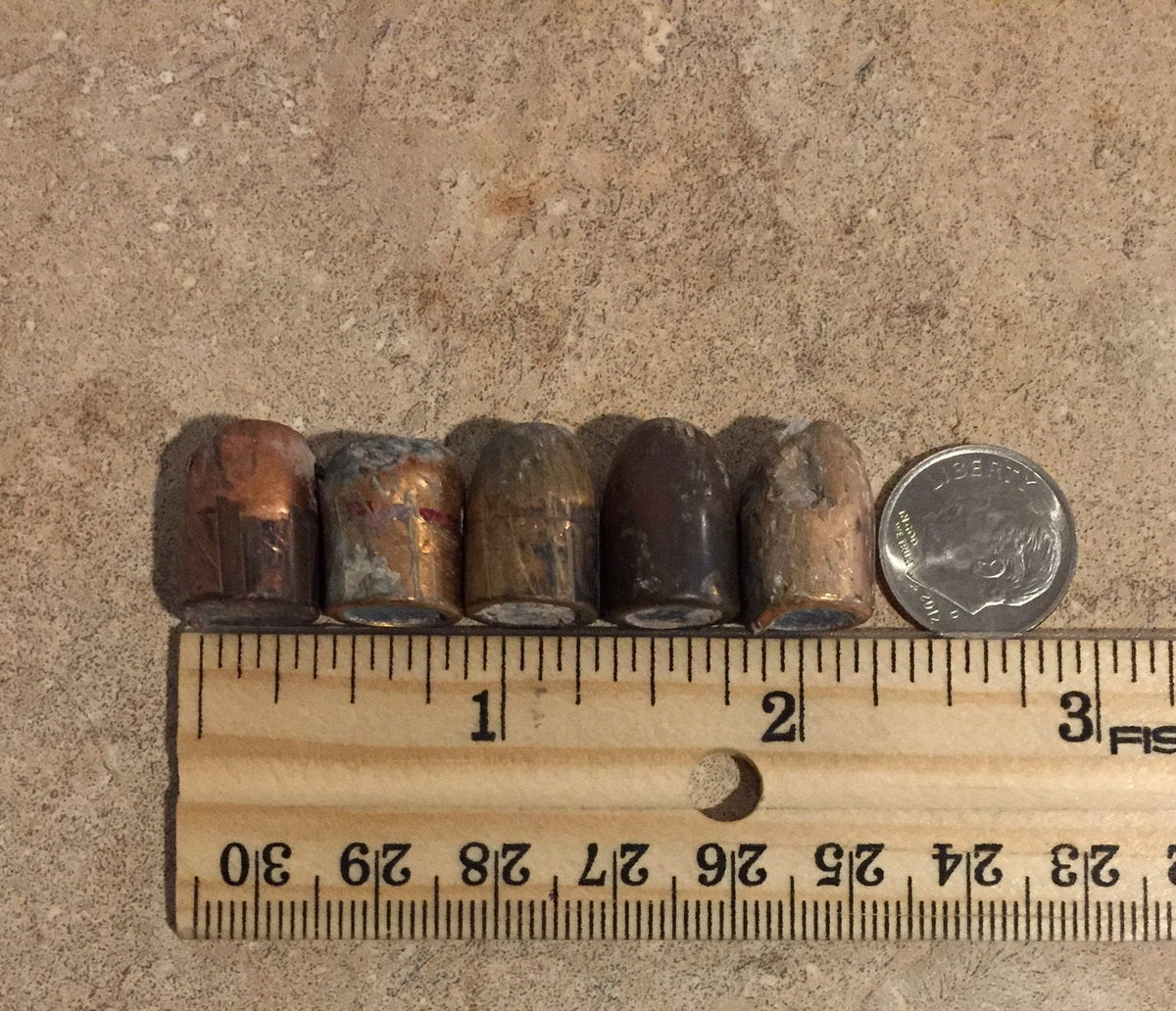 5 tarnished spent bullets lying in a row next to a dime coin and wooden ruler for size comparison
