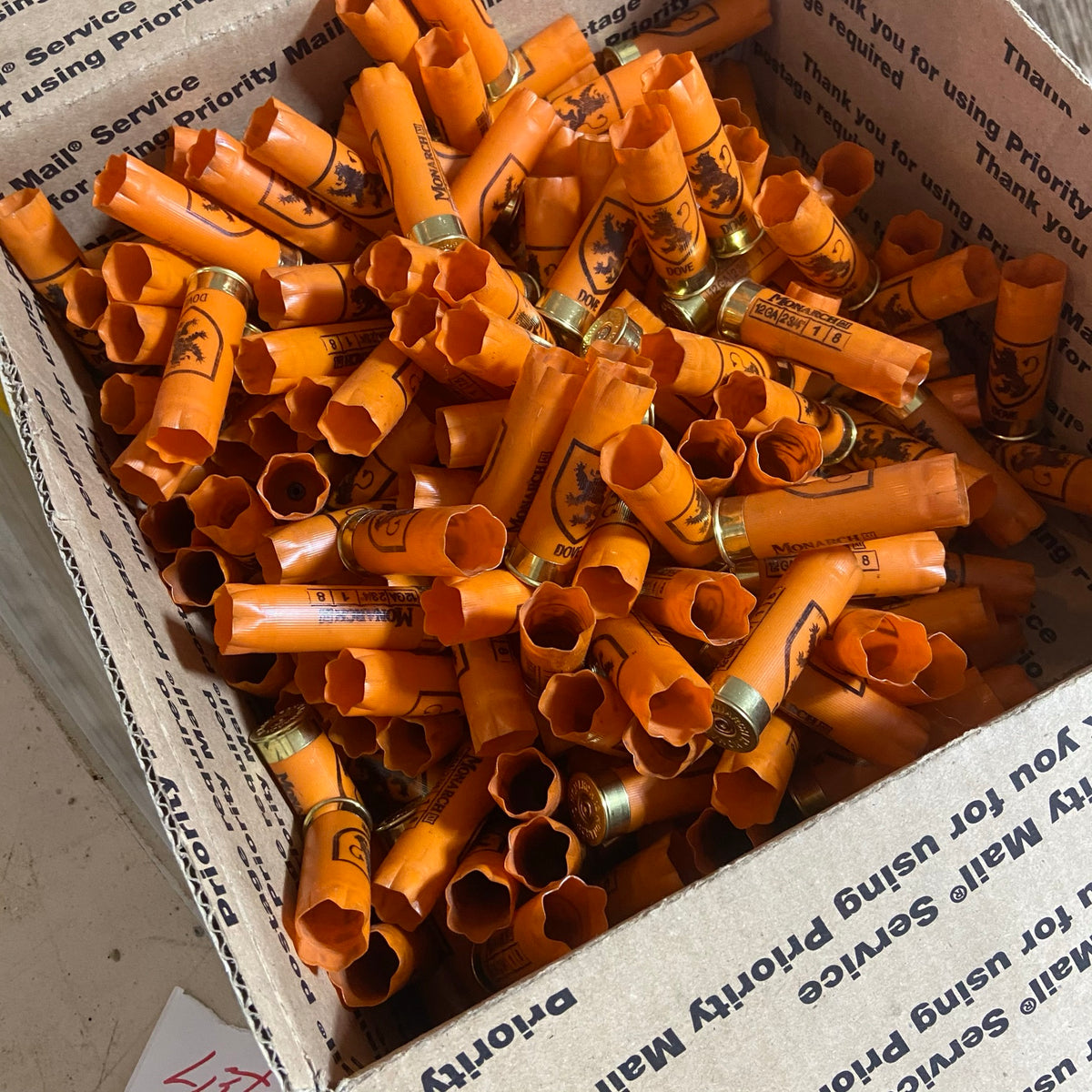 Large pile of empty orange shotgun shells with gold bases in a cardboard box marked Priority Mail
