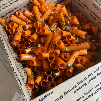 Large pile of empty orange shotgun shells with gold bases in a cardboard box marked Priority Mail