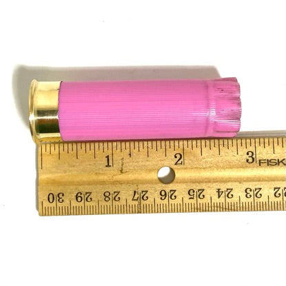 A single empty pink shotgun shell lies against a wooden ruler, measuring approximately 2.5 inches in length.