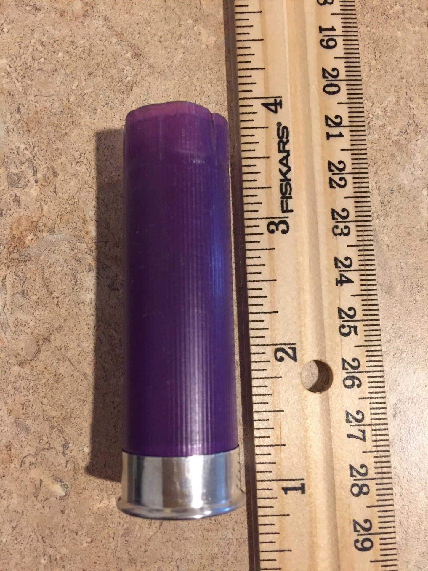 Single purple shotgun shell hull with silver base laid against a wooden ruler measuring about 2.7 inches