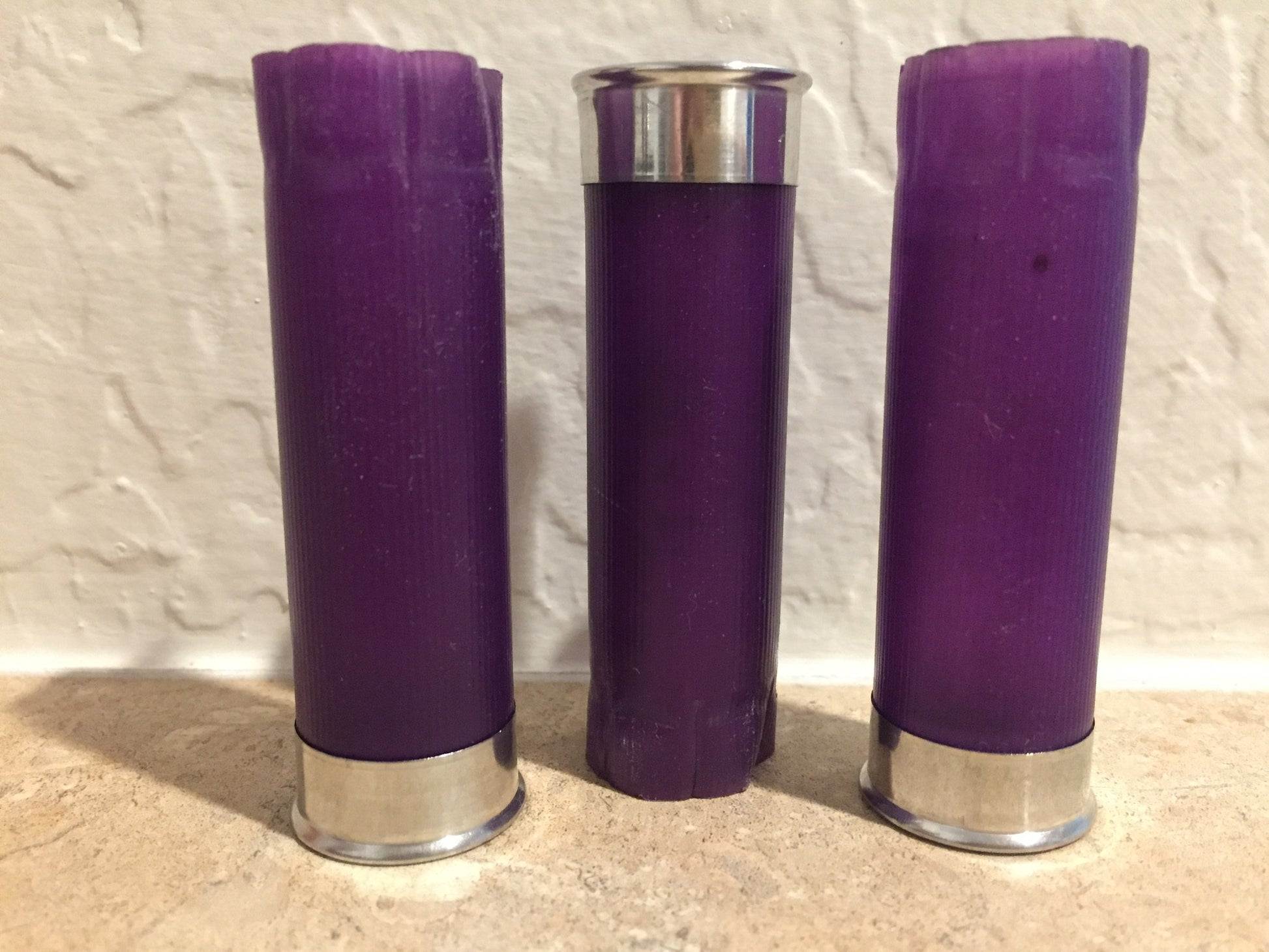 Three purple shotgun shell hulls standing upright on tile against white textured wall, silver bases
