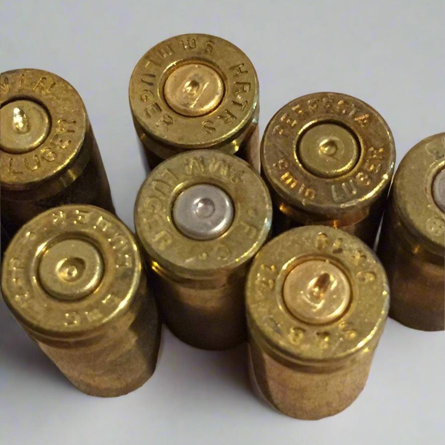 Six raw 9mm brass casings standing upright displaying legible headstamps and indented primers.