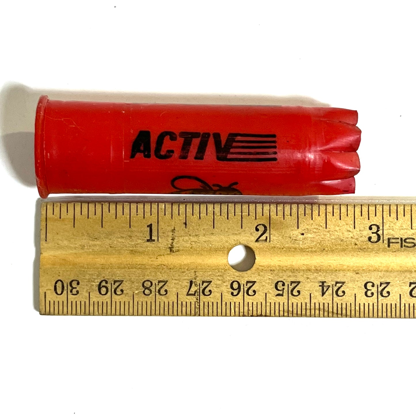 Single red plastic shotgun shell printed "Activ" next to a wooden ruler