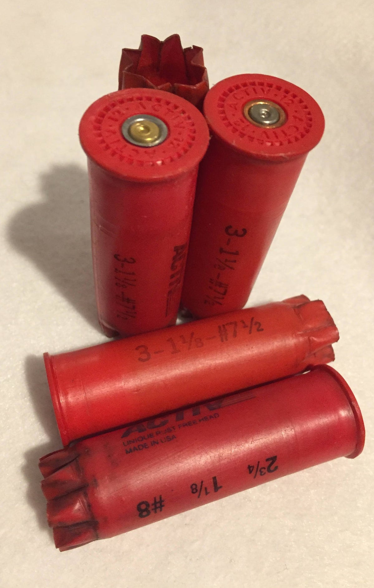 4 red plastic shotgun shells printed "Activ", "3-1⅛-#7½", "2¾ 1⅛ #8" text, 2 standing upright showing brass primers