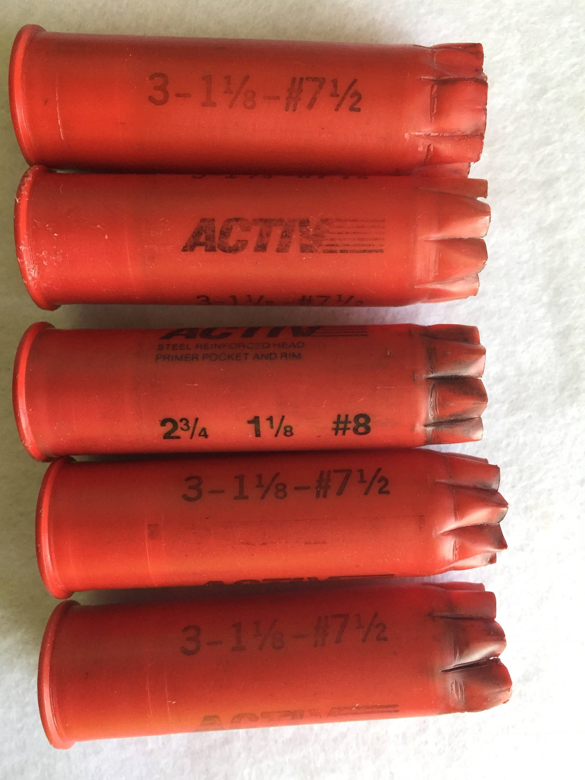 5 red plastic shotgun shells lying in a row printed "Activ", "3-1⅛-#7½", and "2¾ 1⅛ #8" text on white fabric