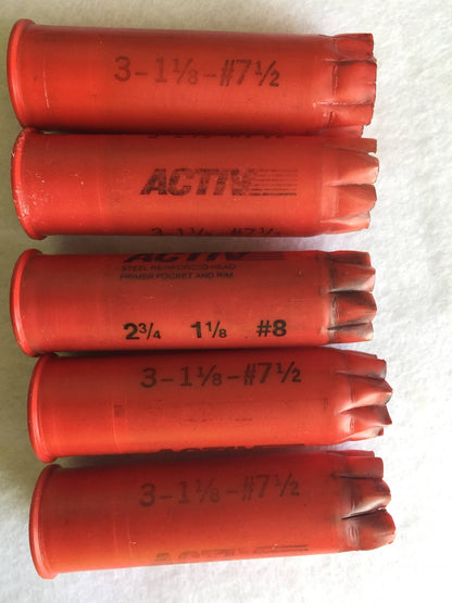 5 red plastic shotgun shells lying in a row printed "Activ", "3-1⅛-#7½", and "2¾ 1⅛ #8" text on white fabric