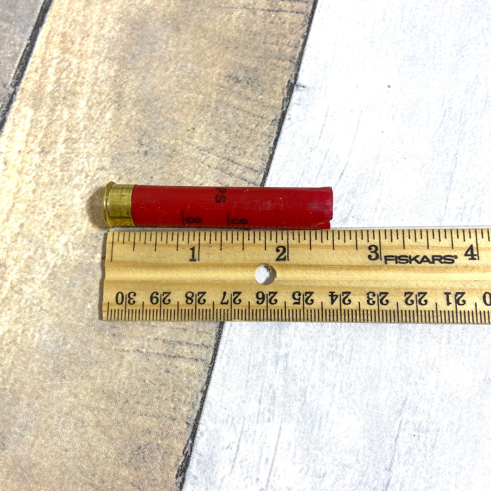 Single empty red 410 bore shotgun shell with brass base next to a wooden ruler showing approximately 2.5 inches in length