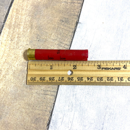 Single empty red 410 bore shotgun shell with brass base next to a wooden ruler showing approximately 2.5 inches in length