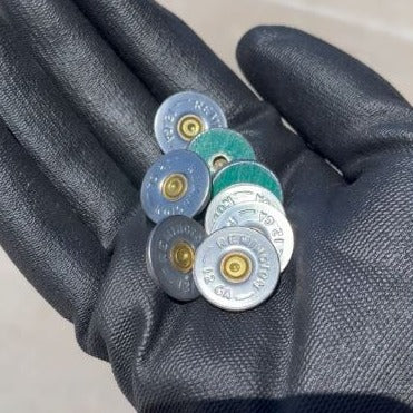 A gloved hand holding five Remington 12 gauge bullet slices to show their size, with the embossed headstamp and one green hull edge visible