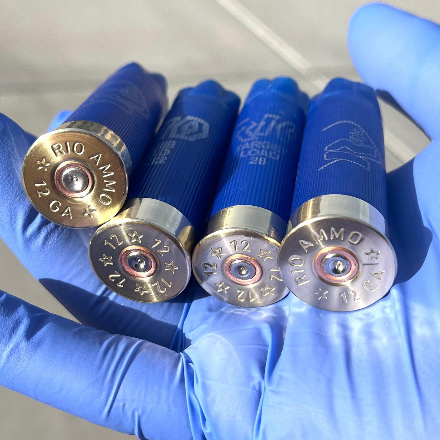 Four blue Rio 12 gauge shotgun shell hulls held in a gloved hand showing brass "RIO Ammo 12 GA" headstamps
