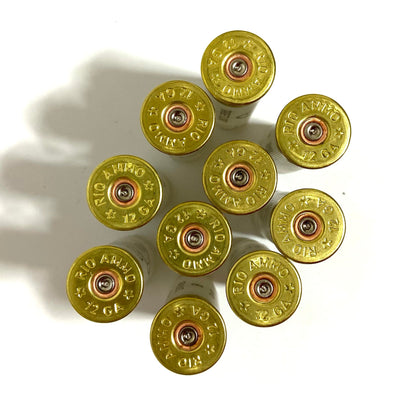Close-up view of multiple gold metal headstamps, spent primers from empty shells showing the Rio 12 GA markings on a light background.
