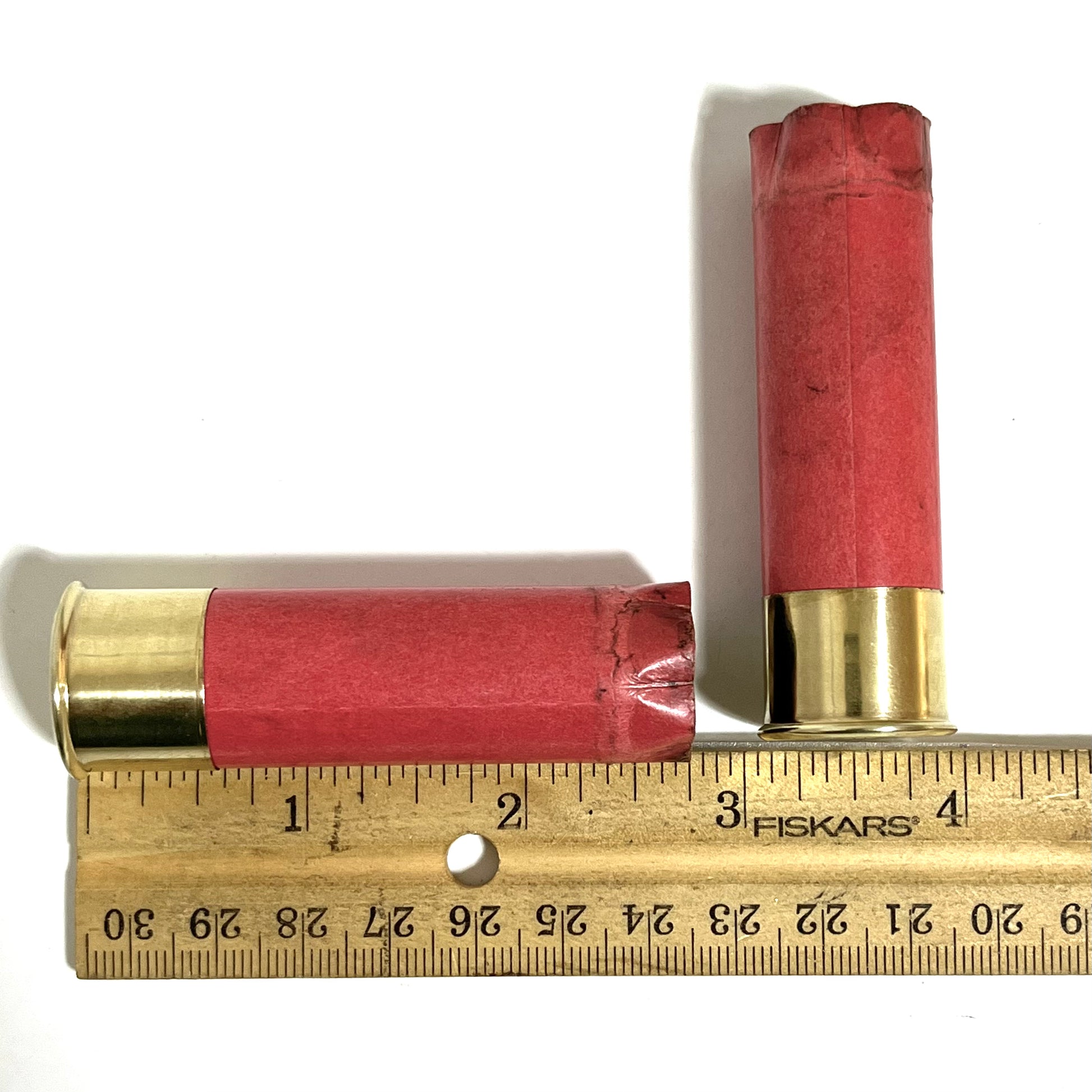 Two salmon pink paper shotgun shells lying next to a wooden ruler showing their length