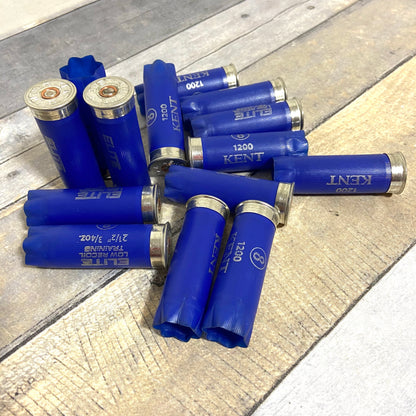 Scattered group of navy blue Kent 12-gauge empty shotgun shells on a weathered wooden background, with some showing silver metal bases and others showing open star crimps.
