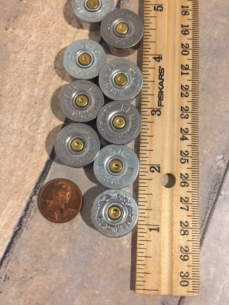 Ten silver 20 gauge shotgun shell brass end caps laid next to a wooden ruler and a penny for size comparison