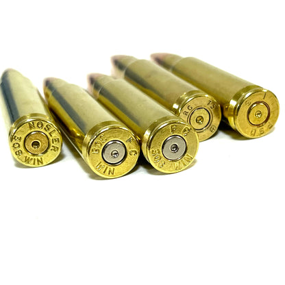 Five brass .308 snap cap props angled to show spent primers and headstamps, including a NOSLER 308 WIN marking
