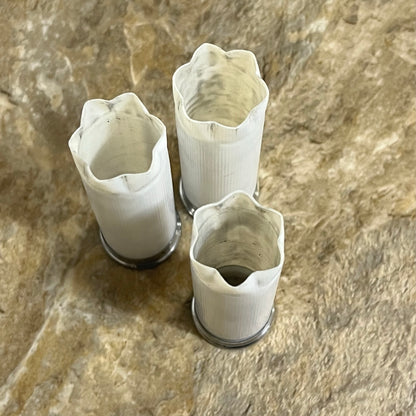 Three white spent shotgun shells standing upright with open frayed tops on a textured tan surface
