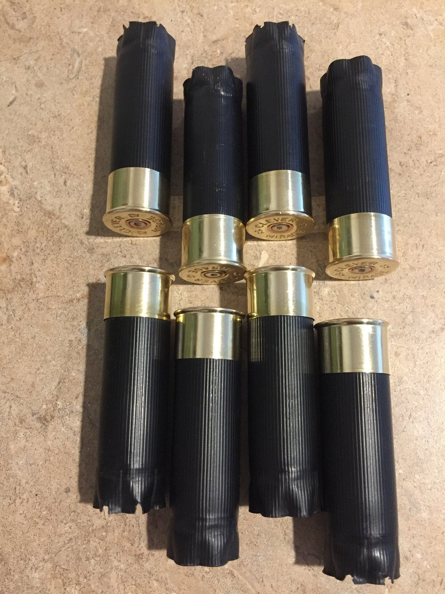 Eight black unmarked shotgun casings arranged in two rows with brass ends facing outward on a tan surface