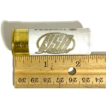 A single white empty dummy round with USA Shooting text laid against a brass ruler showing its length in inches.