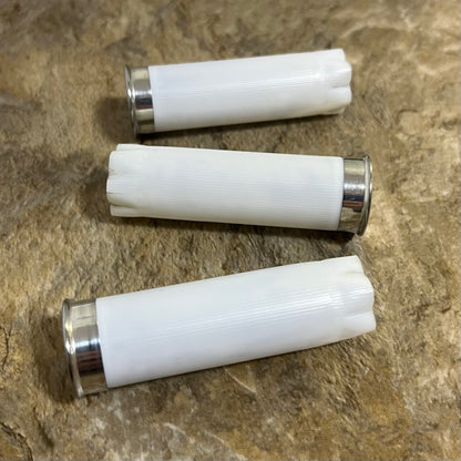 Three white once-fired cartridges with crimped open tops and silver bases resting on a mottled tan surface