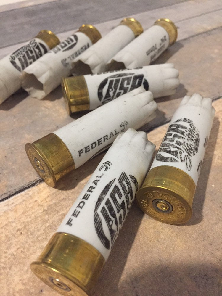 6 white Federal and USA Shooting 12 gauge shotgun shells with brass bases lying on a tile floor