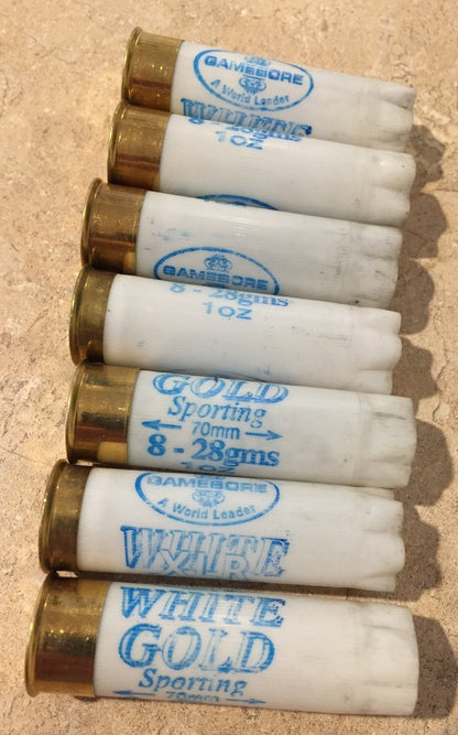 7 white shotgun shells with brass bases lying in a row showing "Gamebore" and "White Gold Sporting" printed text