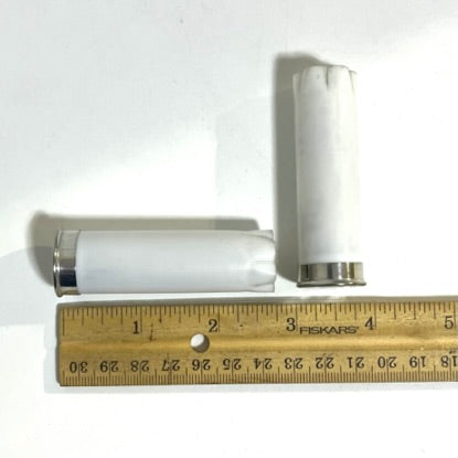 Two white empty shotgun hulls beside a wooden ruler showing about 2 inches, one lying flat and one standing upright