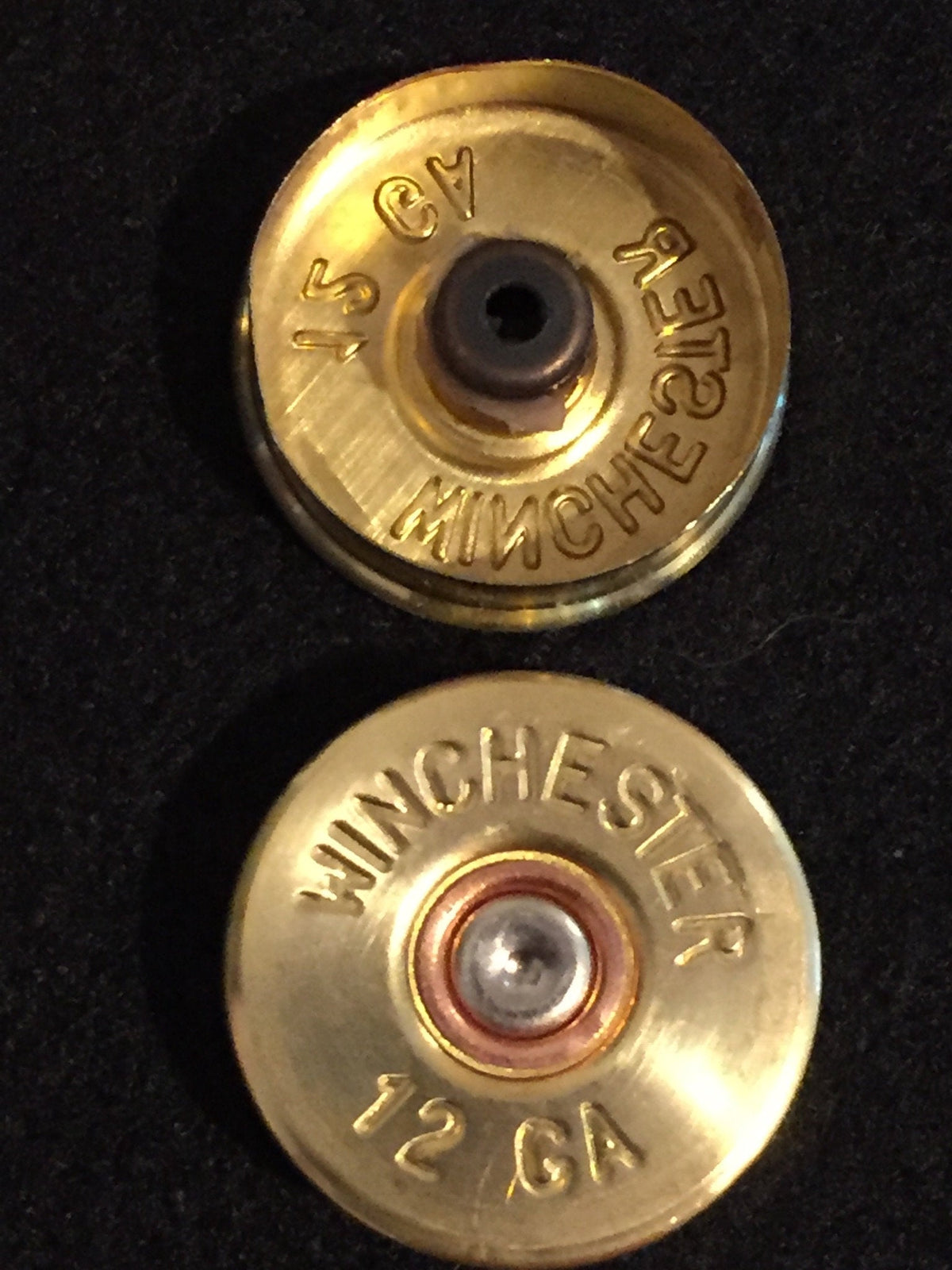2 gold brass shell heads stacked, top showing a hollow primer pocket and bottom showing an intact "Winchester 12 GA" headstamp with silver primer