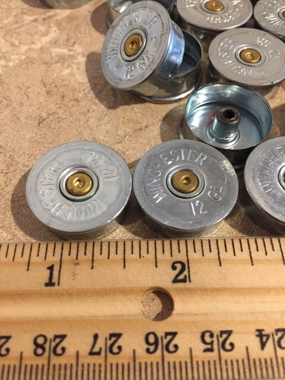 Silver Winchester 12 gauge steel headstamps measured against a ruler on a stone countertop, showing a diameter of about 1.2 inches