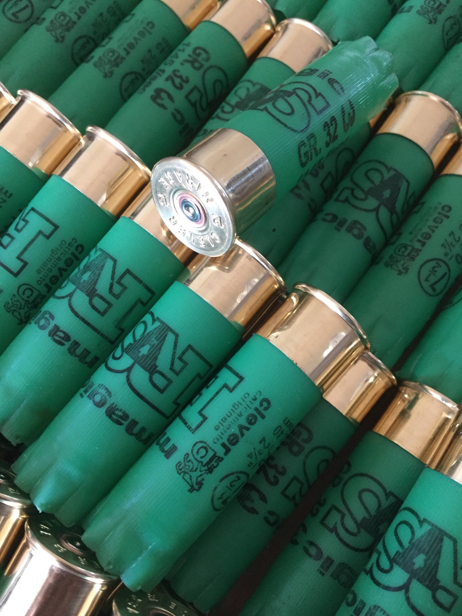 Rows of green shotgun shells with gold bases, one shell tilted showing a silver base with embossed headstamp text