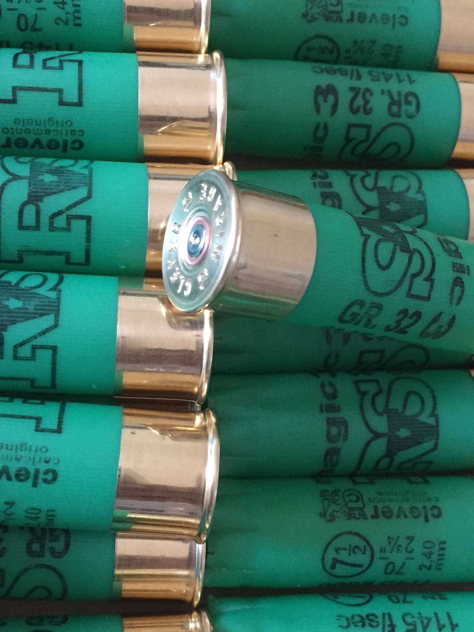 Rows of green shotgun shells with gold bases, one shell tilted showing a silver base with embossed headstamp text